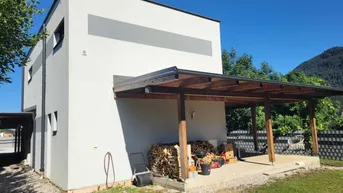 Moderne Hausfassade mit überdachter Terrasse, Holzstapel und Bergblick.