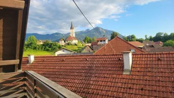 Panoramablick über ein Dorf mit Kirche und Bergen im Hintergrund unter blauem Himmel.