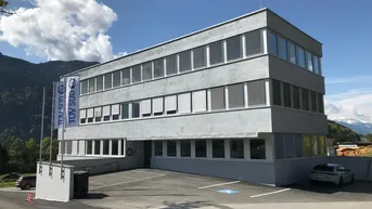 Modernes Bürogebäude mit grauer Fassade, vielen Fenstern und Parkplätzen im Freien vor einer Bergkulisse.