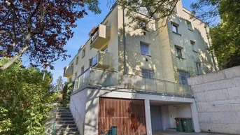 Mehrstöckiges Wohngebäude mit Balkonen, integrierter Garage und Außentreppe, umgeben von Grün.