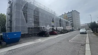 Gebäude unter Gerüst und Schutznetz, das auf Bauarbeiten oder Renovierung hindeutet, mit Straße und parkenden Autos im Vordergrund.