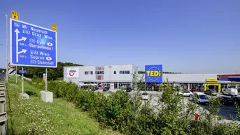 Außenansicht eines Gewerbegebiets mit mehreren Geschäften, Parkplätzen und einer großen blauen Straßentafel.
