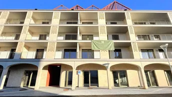 Modernes Mehrfamilienhaus mit mehreren Etagen, Balkonen und Arkaden im Erdgeschoss unter blauem Himmel.