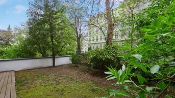 Grüner Gartenbereich mit Rasenfläche und einer kleinen Holzterrasse, umgeben von Bäumen und Gebäuden.
