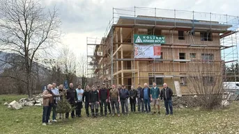 Neubau in Holzbauweise mit Gerüst und einer Gruppe von Personen davor, umgeben von Natur und Bergen.