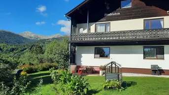 Haus mit Holzbalkon und Bergblick, umgeben von grüner Natur und klarem Himmel.