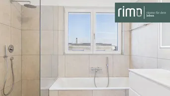 Modernes Badezimmer mit Badewanne, Regendusche und Fenster, hell gefliest und mit Logo.