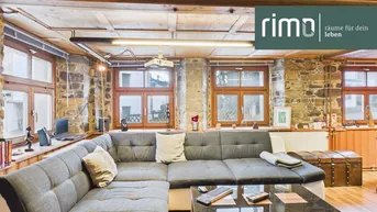 Gemütliches Wohnzimmer mit rustikalen Steinwänden, Holzbalkendecke und großem Ecksofa, umgeben von vielen Fenstern.
