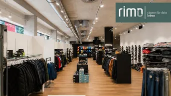 Geräumiger und moderner Verkaufsraum mit Holzboden, Kleiderständern und Schuhregalen unter einer Betondecke.