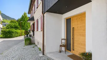 Modernes Haus mit Holztür, gepflastertem Eingangsbereich und Blick auf grüne Hügel.