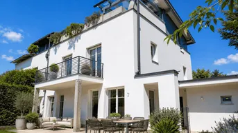 Moderne weiße Villa mit großer Terrasse, Gartenmöbeln und üppiger Bepflanzung unter blauem Himmel.