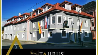 Graues Gebäude mit rotem Ziegeldach und mehreren Dachgauben an einer Straßenecke, mit Werbebanner und Logo.