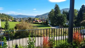Weitläufiger Blick von einem Balkon auf grüne Hügel, ein Tal und ferne Berge unter blauem Himmel.