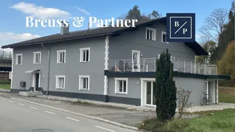 Graues Mehrfamilienhaus mit weiß abgesetzten Fenstern und einem großen Balkon im ersten Stock.