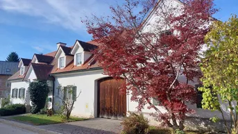 Traditionelles Landhaus mit weißer Fassade, rotem Ziegeldach und herbstlicher Bepflanzung unter blauem Himmel.