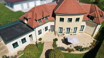 Luftaufnahme eines großen Hauses mit rotem Ziegeldach, weitläufigem Garten und gepflasterter Terrasse.