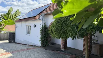 Weißes Haus mit Solaranlage auf dem Dach und gepflastertem Hof, umgeben von grünen Blättern.