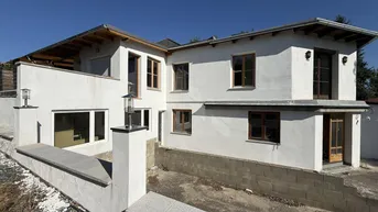 Weiß verputztes Haus mit Holzfenstern und unfertigem Außenbereich unter blauem Himmel.