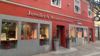 Rote Geschäftsfassade eines Juweliers mit großen Schaufenstern, Eingang und dekorativen Pflanzen am Bürgersteig.