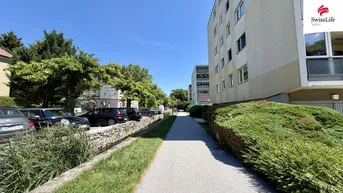 Außenansicht eines Mehrfamilienhauses mit Gehweg, Parkplätzen und grüner Umgebung.
