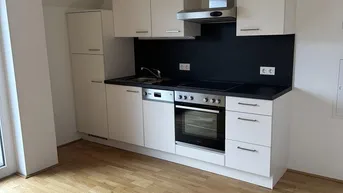 Moderne Einbauküche mit hellen Schränken, dunkler Arbeitsplatte und Holzboden in einem offenen Raum.
