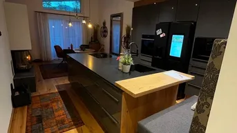 Moderne Küche mit Kochinsel, integrierten Geräten, Holzboden und einem gemütlichen Kamin im Hintergrund.