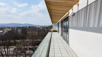 Langer Balkon mit Glasbrüstung und perforiertem Sonnenschutz, bietet weiten Blick über die Stadt.