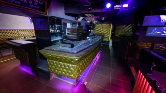 Moderne Bar mit goldgepolsterten Sitzgelegenheiten, glitzernder Theke und violetter Beleuchtung für eine lebendige Atmosphäre.