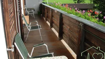 Langer Balkon mit Holzgeländer, Sitzgelegenheiten und Blick auf grüne Wiesen und Berge.