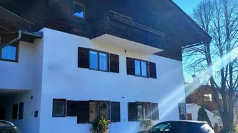 Alpines Gebäude mit weißer Fassade, Holzfensterläden und geparkten Autos davor unter blauem Himmel.
