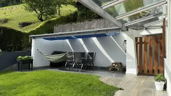 Überdachte Terrasse mit Gartenmöbeln und Hängematte, umgeben von Grünflächen und Hügeln.