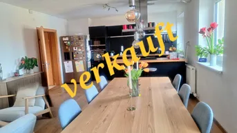 Heller offener Wohn- und Essbereich mit Holztisch, moderner Küche und großen Fenstern.