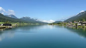 Malerischer See umgeben von Bergen und kleinen Häusern unter blauem Himmel.