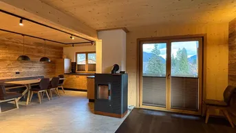 Offener Wohn-Essbereich mit Holzwänden, Kaminofen und Zugang zum Balkon.