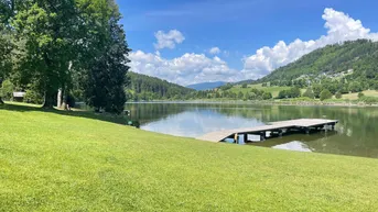 Idyllische Seenlandschaft mit Holzsteg, umgeben von grünen Wiesen und bewaldeten Hügeln unter blauem Himmel.