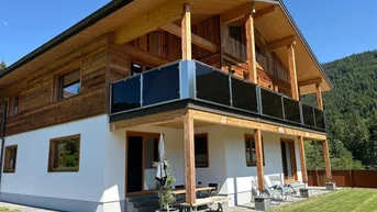 Modernes Holzhaus mit Balkon, Terrasse und gepflegtem Rasen in alpiner Umgebung.