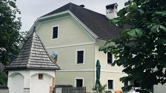 Traditionelles Gebäude mit hellgelber Fassade, umgeben von Grünflächen und einem kleinen Schrein.