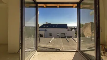 Blick aus einem Fenster auf ein Flachdach und ein angrenzendes Gebäude unter blauem Himmel.