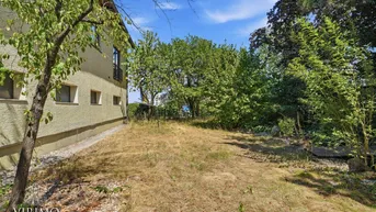 Seitenansicht des Gebäudes mit einem trockenen Gartenbereich und altem Baumbestand unter blauem Himmel.