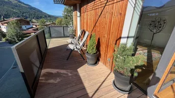 Sonniger Balkon mit Holzboden, Glasgeländer und Blick auf die umliegende Berglandschaft.