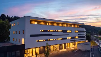 Modernes Gebäude bei Sonnenuntergang mit beleuchteter Fassade und umliegender Natur.