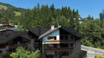 Mehrfamilienhaus mit mehreren Balkonen und grüner Umgebung, umgeben von Bäumen und Hügeln.