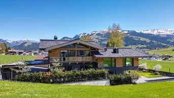 Modernes Chalet-Haus mit Holz- und Steinfassade, umgeben von grünen Wiesen und Bergen.