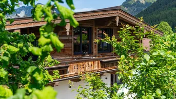 Traditionelles Holzhaus im alpinen Stil, umgeben von üppigem Grün und Bergen im Hintergrund.