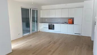 Heller offener Wohnbereich mit integrierter weißer Küche, Parkettboden und Zugang zum Balkon.