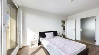 Helles Schlafzimmer mit Holzboden, großem Fenster und Zugang zu einem weiteren Raum.