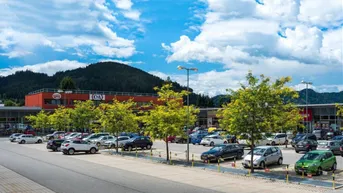 Einkaufszentrum mit großem Parkplatz und Geschäften unter blauem Himmel und Bergen im Hintergrund.