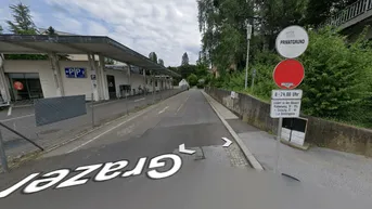 Blick auf eine Straße mit angrenzendem Privatgrundstück, Parkplatz und Bäumen unter bewölktem Himmel.