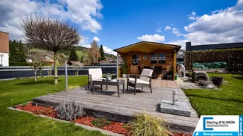 Gepflegter Garten mit Holzterrasse, Gartenhaus und Sitzgelegenheiten unter blauem Himmel.