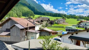 Malerischer Ausblick auf ein Bergdorf mit grünen Wiesen und majestätischen Bergen unter blauem Himmel.
